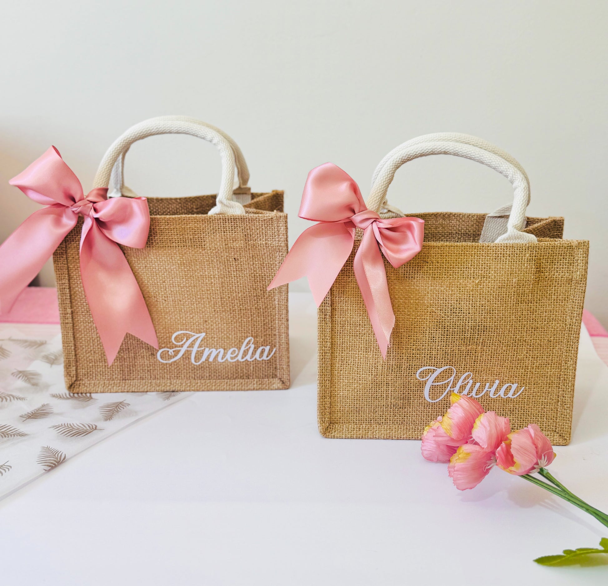 Two small jute bags with pink ribbons and names 'Amelia' and 'Olivia' on a light background.